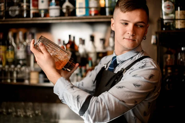 Young handsome bartender standing behind the bar counter with glassy shaker.