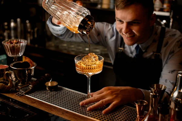 Young smiling bartender pours finished cold cocktail from glassy shaker into glass. — Stock Photo, Image