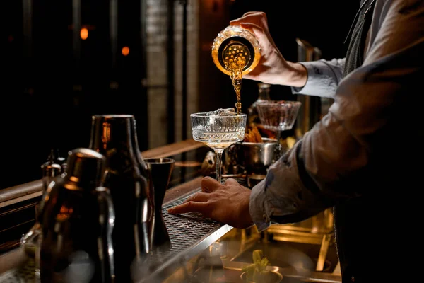 Young bartender professionally pours finished cold brown cocktail from glass shaker into wineglass. — Stock Photo, Image