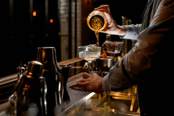 Young bartender professionally pours finished cold brown cocktail from glass shaker into wineglass.