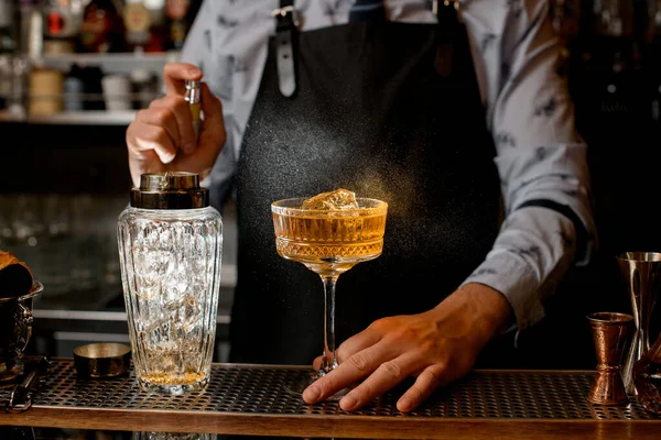 Barman en delantal negro espolvorea sobre vidrio con cóctel . — Foto de Stock