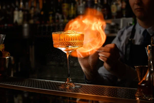 Glass of alcoholic drink and ice stands on bar counter. Bartender makes fire flame behind it. — Stock Photo, Image