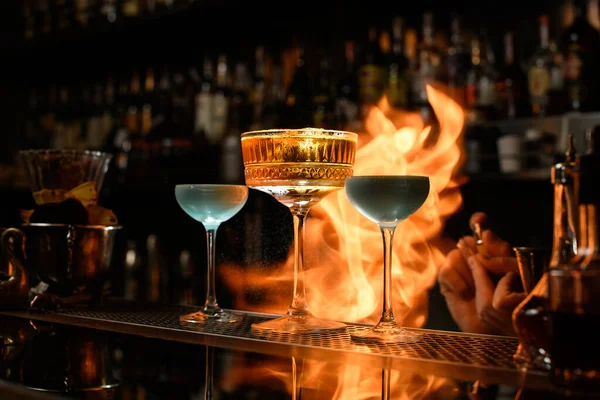 Several glasses of alcoholic drinks stands on bar counter. Fire flame behind it. — Stock Photo, Image