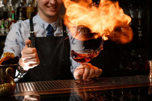 Jovem garçom sorrindo segura vidro com coquetel, pulverizando sobre ele e incendeia . — Fotografia de Stock