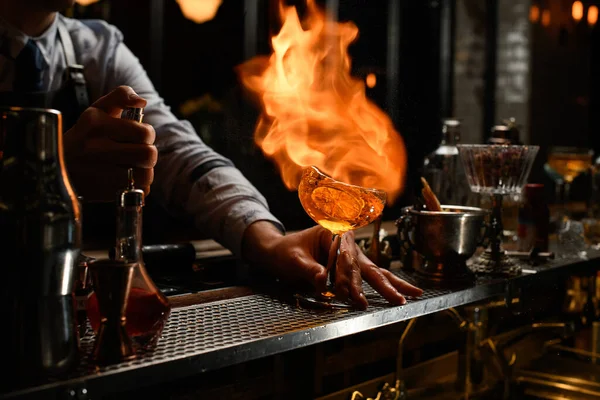 Bartender holds glass with cocktail, spraying on it and sets on fire. — Stock Photo, Image