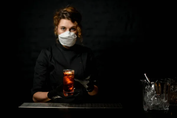 Mulher jovem barman em máscara médica detém vidro com bebida fria em suas mãos — Fotografia de Stock