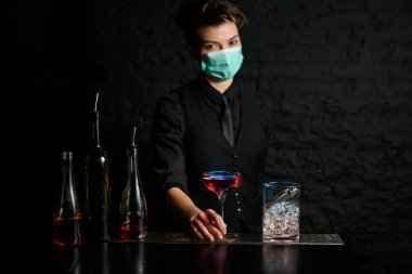 Barda duran siyah takım elbiseli ve tıbbi maskeli bir barmen..