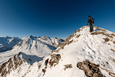 Kayak ekipmanlı bir adam dağa tırmanır ve başka bir sporcu yükselir..