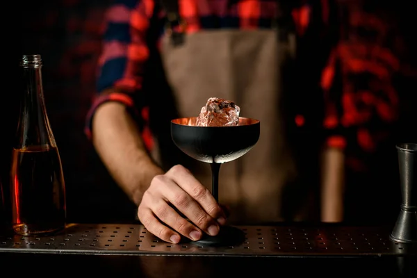 Close-up. Barmans hand holds black frosted glass with piece of ice — Stock Photo, Image