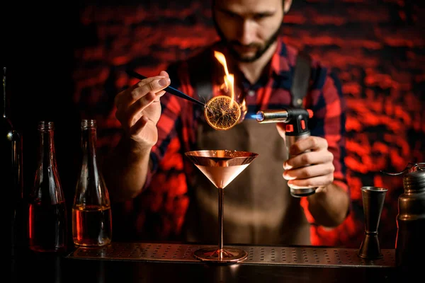 Hombre en el bar decorado con vidrio de martini de metal por corte de cítricos y lo prende fuego — Foto de Stock