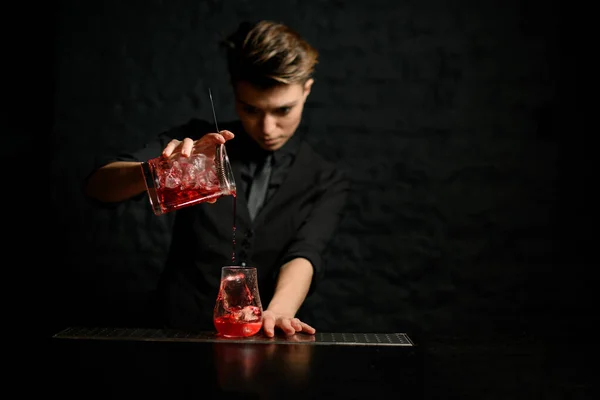 Ung bartender häller försiktigt cocktail från att blanda kopp med sil i glas is — Stockfoto