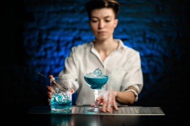 Genç bayan barmen elinde mavi cam bardakla barın üstünde mavi soğuk kokteyl standı tutuyor.
