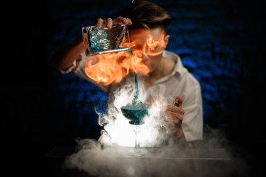 Kadın barmen, dumanlı barda bardağa karıştırdığı bardaktan mavi kokteyl döker ve üzerine ateş püskürtür.
