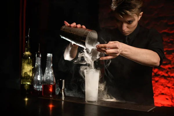 Camarero profesional en bar oscuro vierte suavemente cóctel de agitador de acero en vidrio ahumado — Foto de Stock