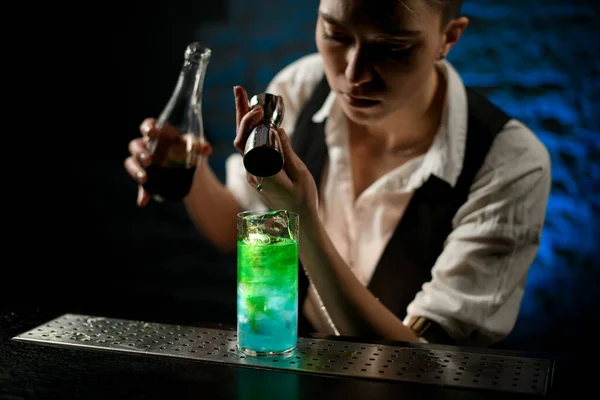 Young professional bartender adds green drink into glass on bar counter.