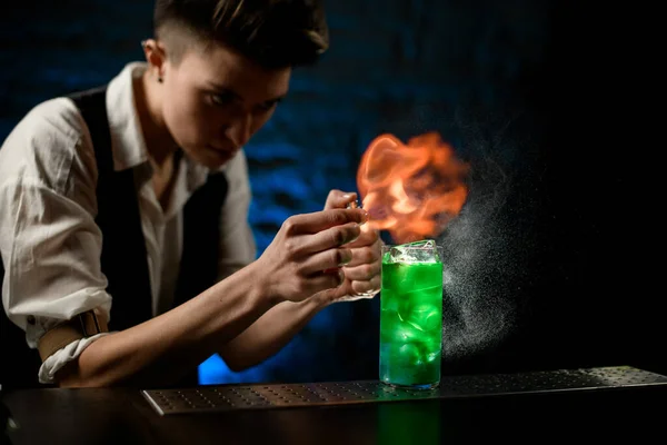 Camarero espolvorea sobre vidrio con cóctel verde brillante y hace llama de fuego sobre él . — Foto de Stock