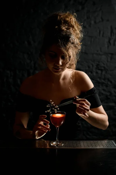 Beautiful young woman at bar decorates glass with alcoholic drink by flowering branch