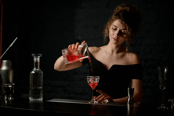 Mulher atraente bartender em blusa preta derrama bebida alcoólica em vidro no bar . — Fotografia de Stock