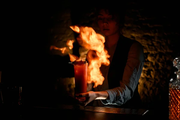 Camarera mujer sosteniendo vidrio con cócteles espolvorea sobre él y magistralmente hacer fuego . — Foto de Stock