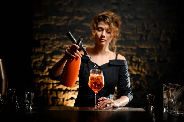 Young bartender girl attentively pours carbonated drink to glass with negroni cocktail.