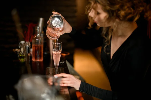 bartender lady attentively pours ready-made cold cocktail from mixing cup into glass