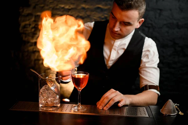 Primo piano. Barman spruzza attivamente su vetro con cocktail e fa fuoco . — Foto Stock