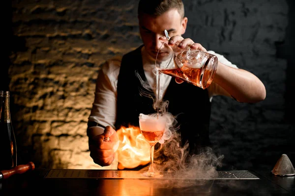 Primo piano. Il barman versa con attenzione il cocktail a spruzzi di vetro fumosi e accende il fuoco . — Foto Stock