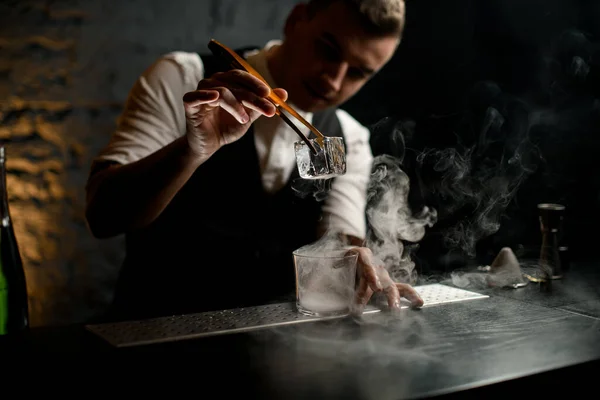 Homem barman suavemente segura na mão pinças com gelo sobre vidro fumegante e olhar para ele . — Fotografia de Stock