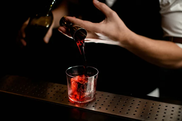 Primer plano. Barmans mano vierte bebida roja de jigger en vidrio con hielo —  Fotos de Stock