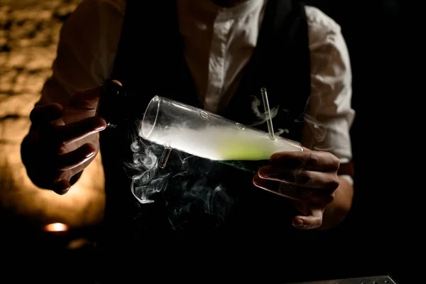 Man bartender at dark bar holds in his hands illuminated designer glass flask with smoky canabis cocktail inside