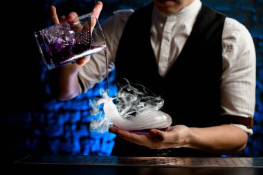 Yakın plan barmen cam karıştırıcı bardaktan içeceği elindeki sıra dışı şekilli dumanlı cam şişeye ustaca dolduruyor.