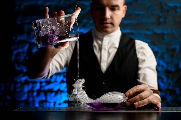 Κοντινό πλάνο μπάρμαν προσεκτικά ρίχνει ποτό από το ποτήρι ανάμιξης γυαλί σε καπνιστή γυάλινη φιάλη ασυνήθιστου σχήματος — Φωτογραφία Αρχείου