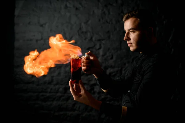Barman profesional sostiene sus manos vaso con cóctel y hacer fuego llama sobre él . — Foto de Stock