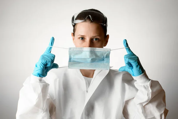 Femme en costume de protection blanc et gants tient un masque médical dans ses mains . — Photo