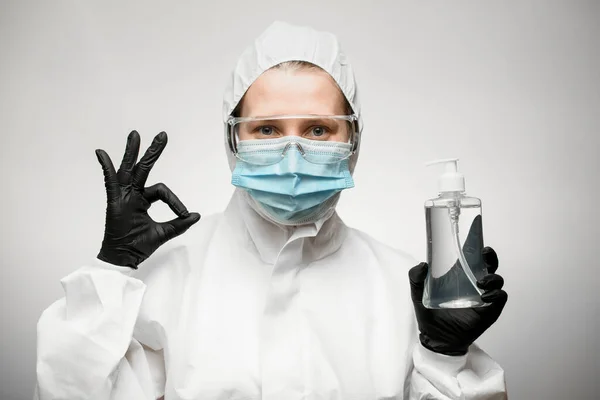 Γυναίκα σε ιατρική μάσκα, γυαλιά και λευκό προστατευτικό κοστούμι δείχνει χέρι εντάξει σημάδι. — Φωτογραφία Αρχείου