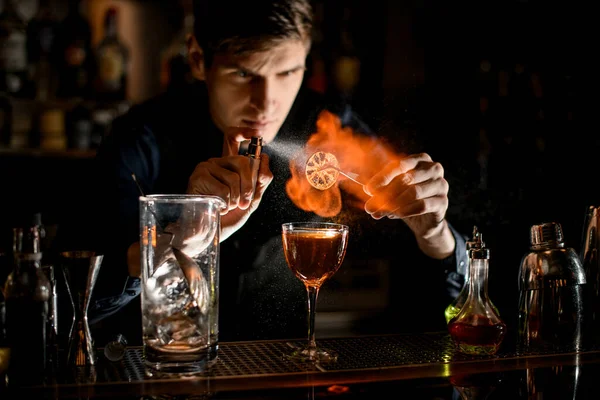 Mladý barman drží pinzetu s plátkem citrusu nad skleněnými postřikovači a zapaluje ji. — Stock fotografie