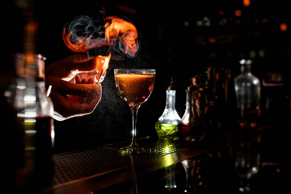 Barman mano espolvorear a cóctel alcohólico con trozo de ralladura de cítricos y hacer fuego — Foto de Stock