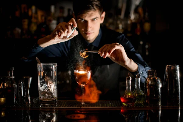 Профессиональный бармен держит пинцет с кусочком цитрусовых над стеклянными брызгами и поджигает его . — стоковое фото