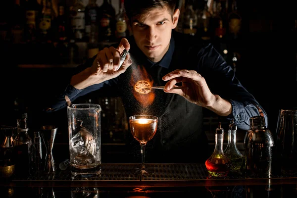 Genç barmen profesyonel olarak cımbızla cam parçalarının üzerine narenciye serpiştirir ve ateşe verir.. — Stok fotoğraf