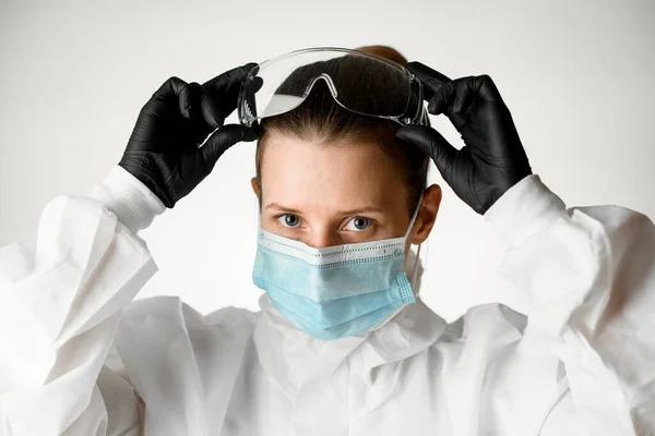 Enfermera joven en máscara médica y traje protector blanco ajusta las gafas en la cabeza —  Fotos de Stock
