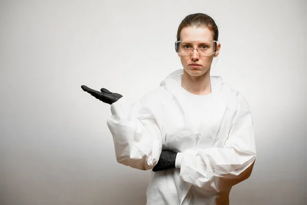 Young woman in full medical protective clothing stands on grey background and shows hand to side — Stock Photo, Image