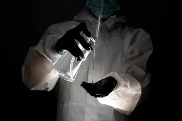 Человек в защитной одежде держит бутылку дезинфицирующего средства и наливает его на руку в темной комнате — стоковое фото