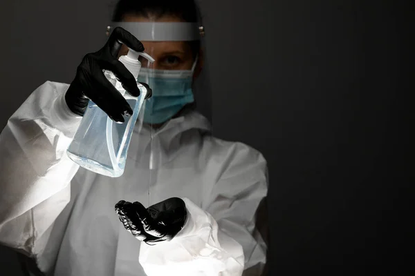 Πρόσωπο με προστατευτική ενδυμασία κρατά ένα μπουκάλι απολυμαντικό και το ρίχνει στο χέρι στο σκοτεινό δωμάτιο — Φωτογραφία Αρχείου