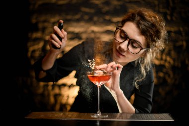 Güzel genç bir barmen dikkatle bir bardak içkiyi süsler ve üzerine serper.