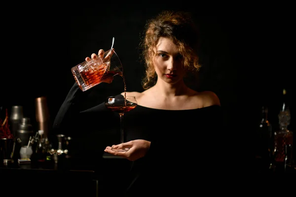 Jovem bartender mulher em blusa preta segura vidro e derrama coquetel nele . — Fotografia de Stock