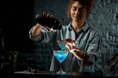 Bardaki kız bardaktan bardağa parlak mavi bir içki döker.