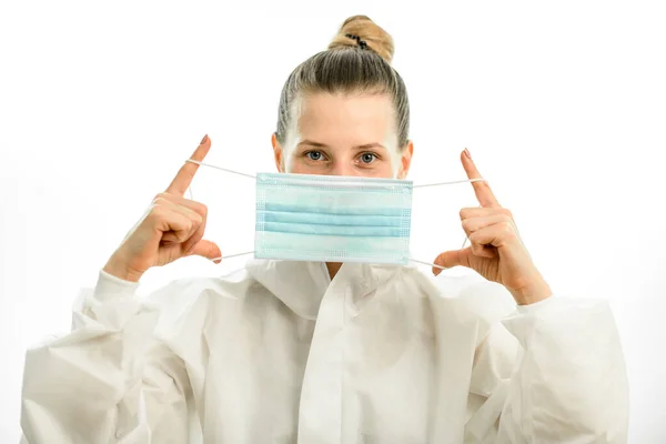 Wanita berambut pirang dalam setelan pelindung putih memegang topeng medis di depan wajahnya — Stok Foto