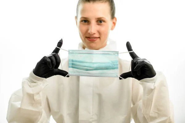 Молодая женщина в белом защитном костюме и черных перчатках держит медицинскую маску в руках — стоковое фото