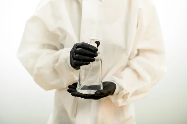 Close-up van de fles met ontsmettingsmiddel in handen van persoon in zwarte handschoenen en wit beschermingspak — Stockfoto