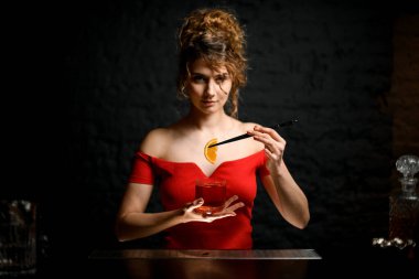 Güzel kadın profesyonel barmen bardağı kokteyl ile tutuyor ve onu portakal dilimiyle süslüyor..